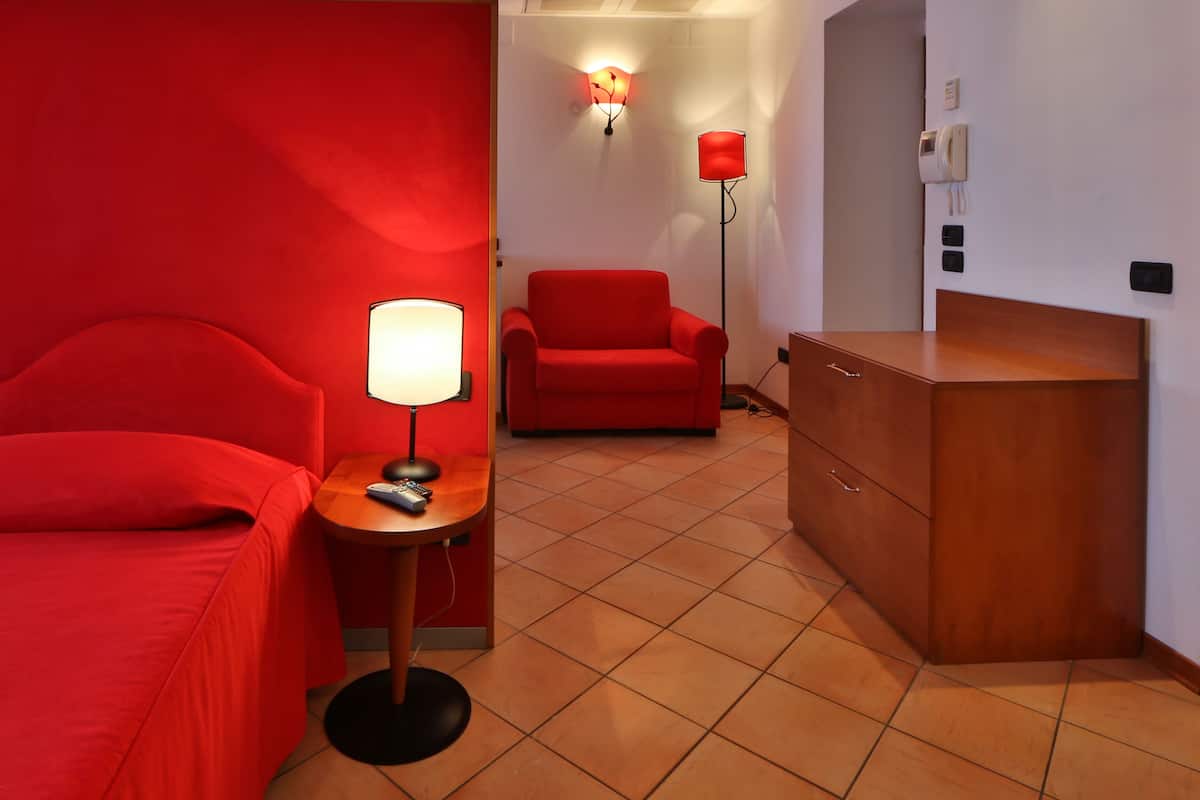Une chambre d’hôtel avec un lit rouge, un fauteuil rouge, une petite table avec une lampe et une commode en bois.