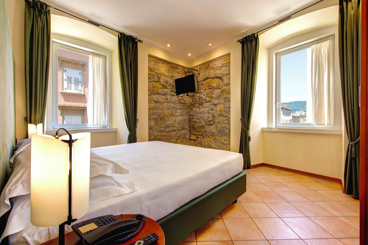 A hotel room with a bed, a lamp, a telephone, a stone wall, and a TV mounted on the wall.
