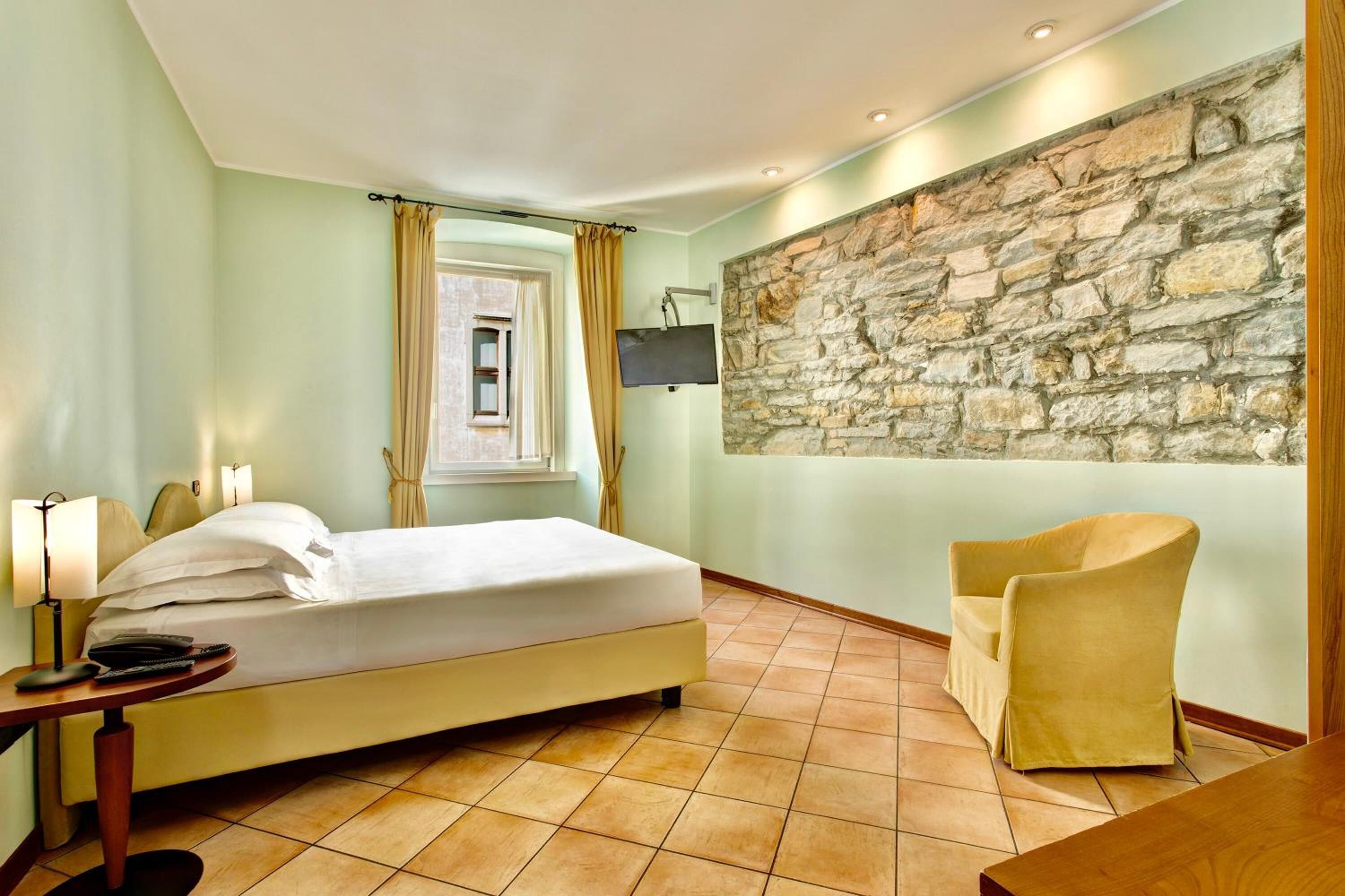 A hotel room with a bed, a chair, a TV mounted on a stone wall, and a window with curtains.
