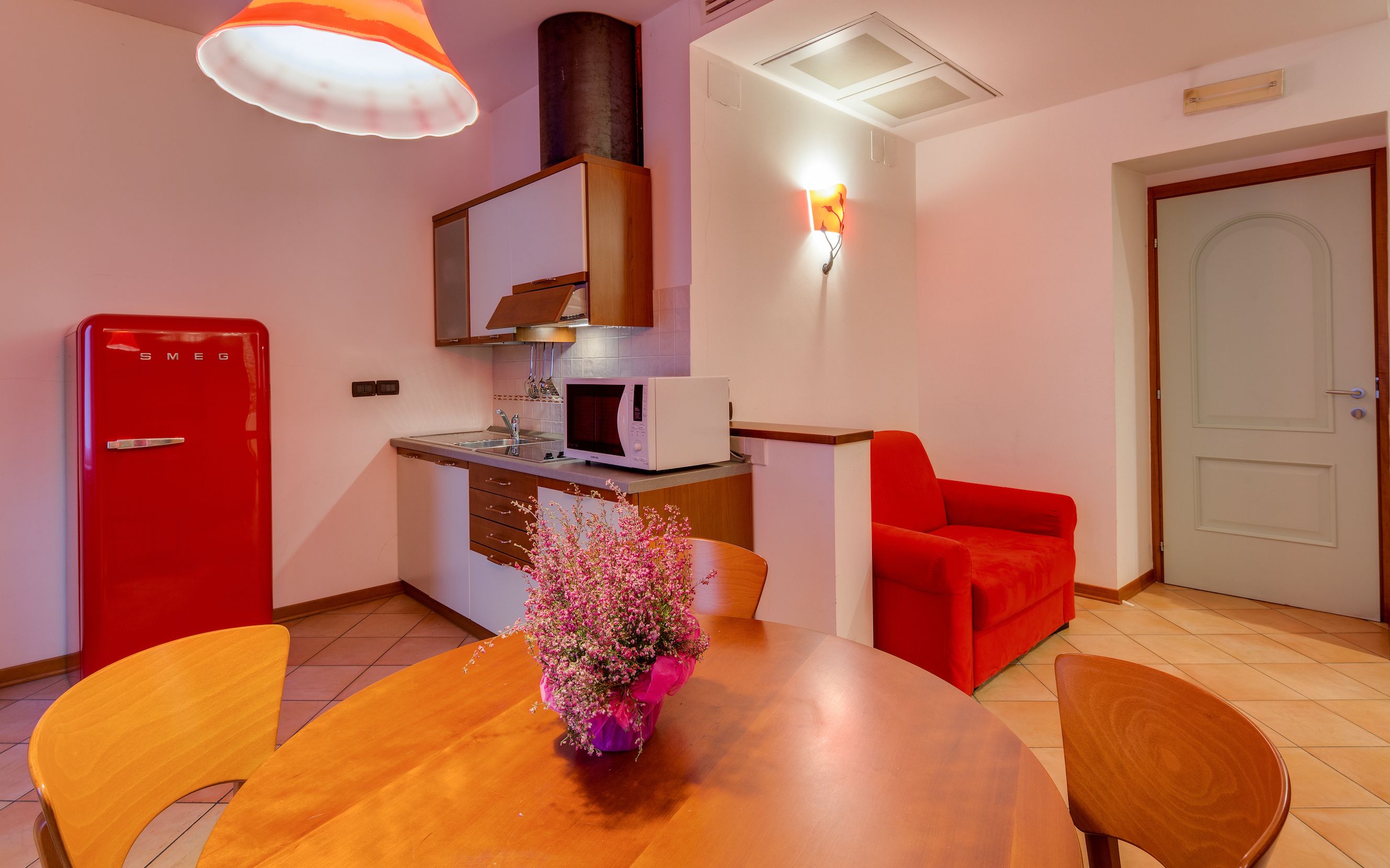 A compact kitchen with a red refrigerator, a microwave, and a sink. There's a wooden dining table with chairs and a vase of flowers. A red armchair is against the wall, and a white door is visible.
