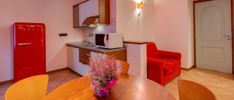 A compact kitchen with a red refrigerator, a microwave, and a sink. There's a wooden dining table with chairs and a vase of flowers. A red armchair is against the wall, and a white door is visible.