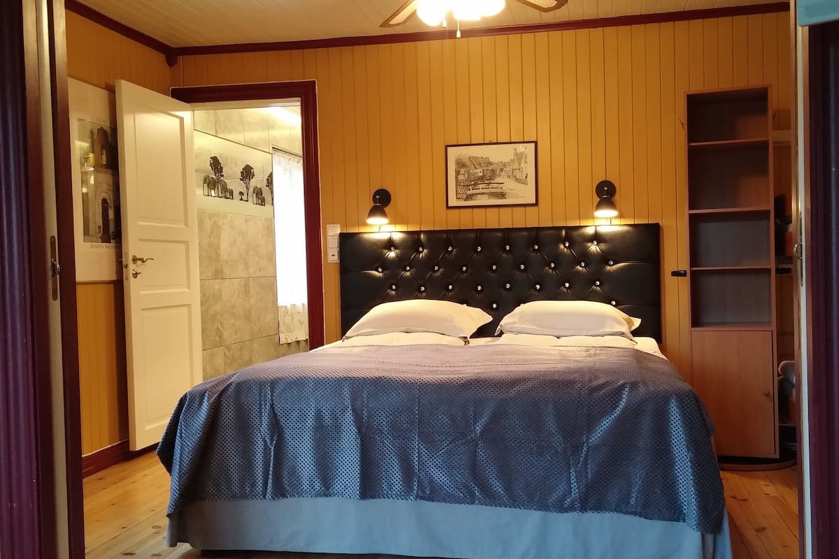 A bedroom with a large bed, wooden panel walls, two wall-mounted lamps, and a framed picture above the bed.