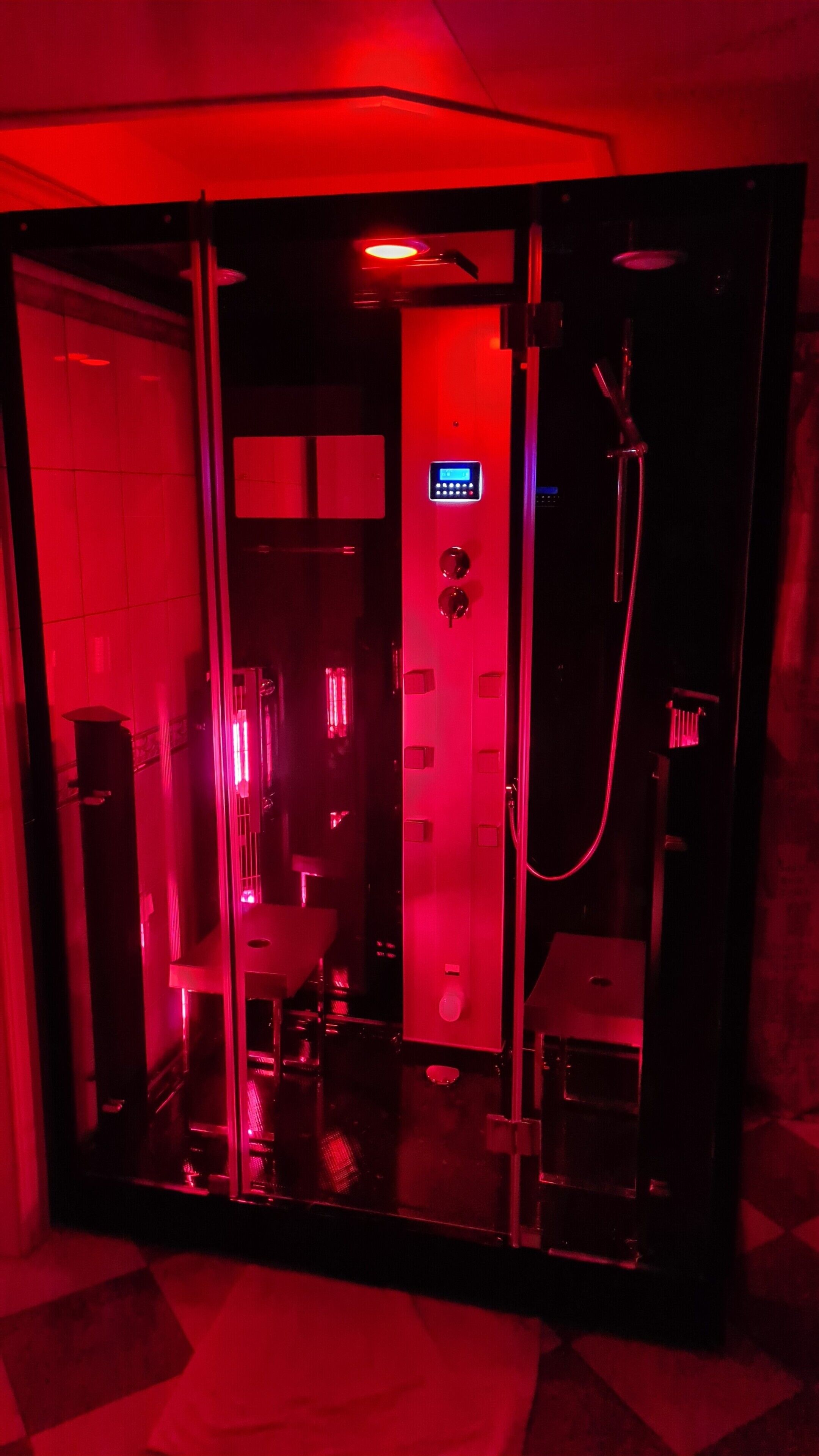 Un baño con iluminación roja, cabina de vidrio, panel de control digital y zona de ducha.