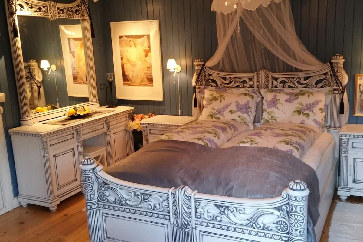A bedroom with a four-poster bed, a vanity with a mirror, and a nightstand with a lamp.