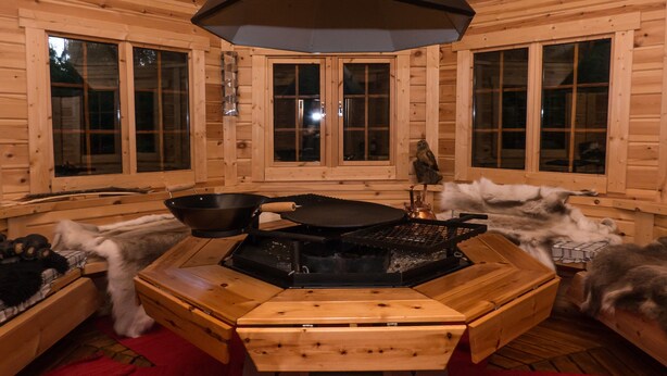 A wooden cabin interior with a central cooking area featuring a stove, a frying pan, and a kettle. There are wooden cabinets and a window with a view of the outdoors. - Solheim Overnatting (Midt-Telemark)