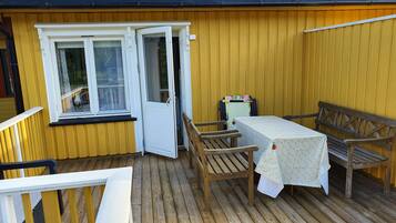 Una terraza de madera con una mesa y sillas, una mesita con mantel floreado, y una puerta que da a una habitación.
