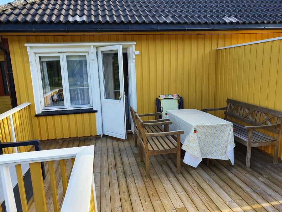 Una terraza de madera con una mesa y sillas, una mesita con mantel floreado, y una puerta que da a una habitación.
