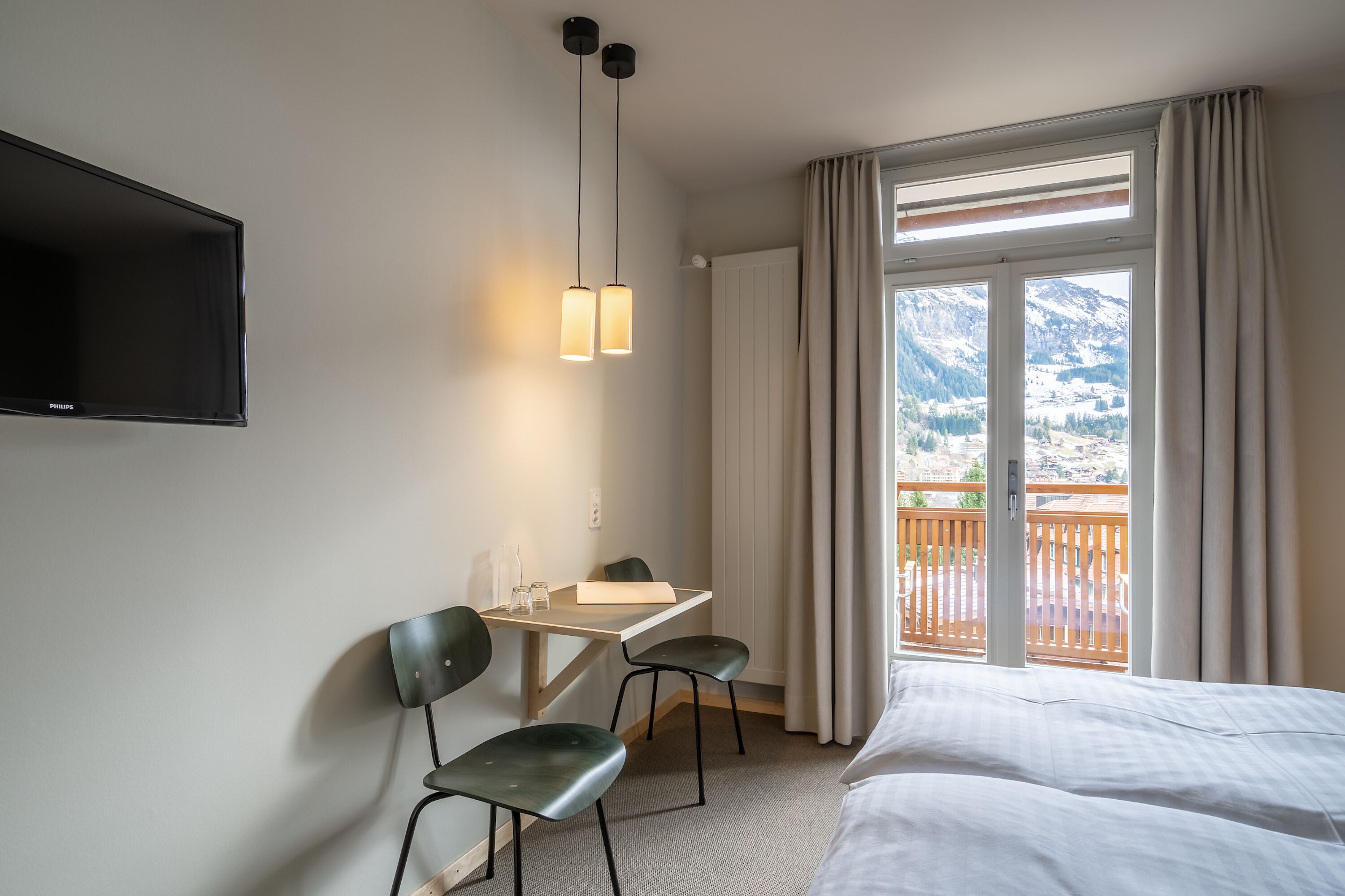 A hotel room with a bed, a desk with two chairs, a flat-screen TV, and a balcony with a mountain view.
