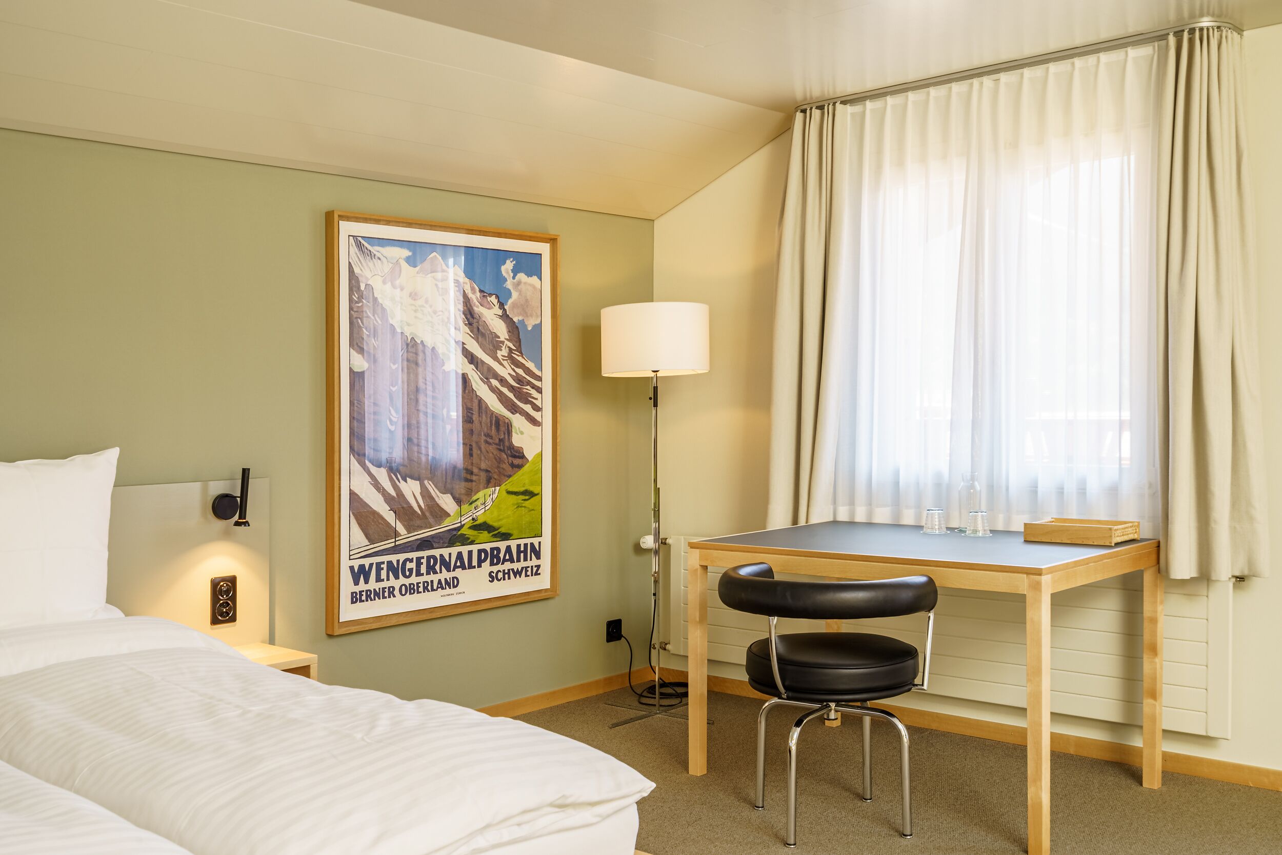 A hotel room with a bed, a desk with a chair, a lamp, and a poster on the wall.