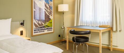 A hotel room with a bed, a desk with a chair, a lamp, and a poster on the wall.
