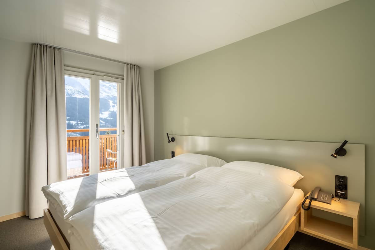 Ein Hotelzimmer mit einem großen Bett, einem Nachttisch mit Lampe und Blick auf Berge durch das Fenster.