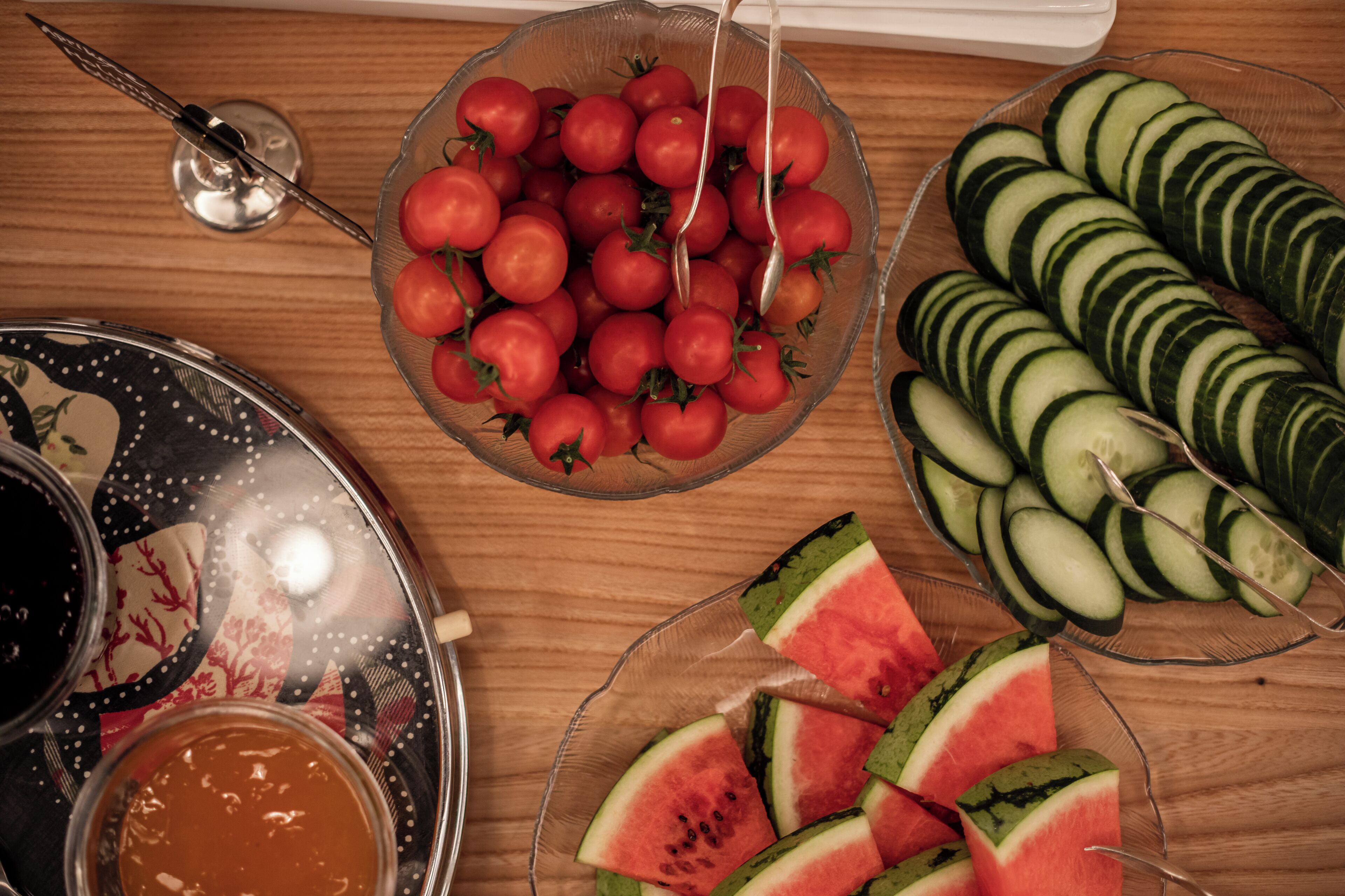 A wooden table with a glass bowl of cherry tomatoes, a plate of sliced cucumbers, and a bowl of watermelon slices.
