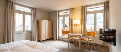 A hotel room with a bed, a desk, a chair, a TV, and large windows.