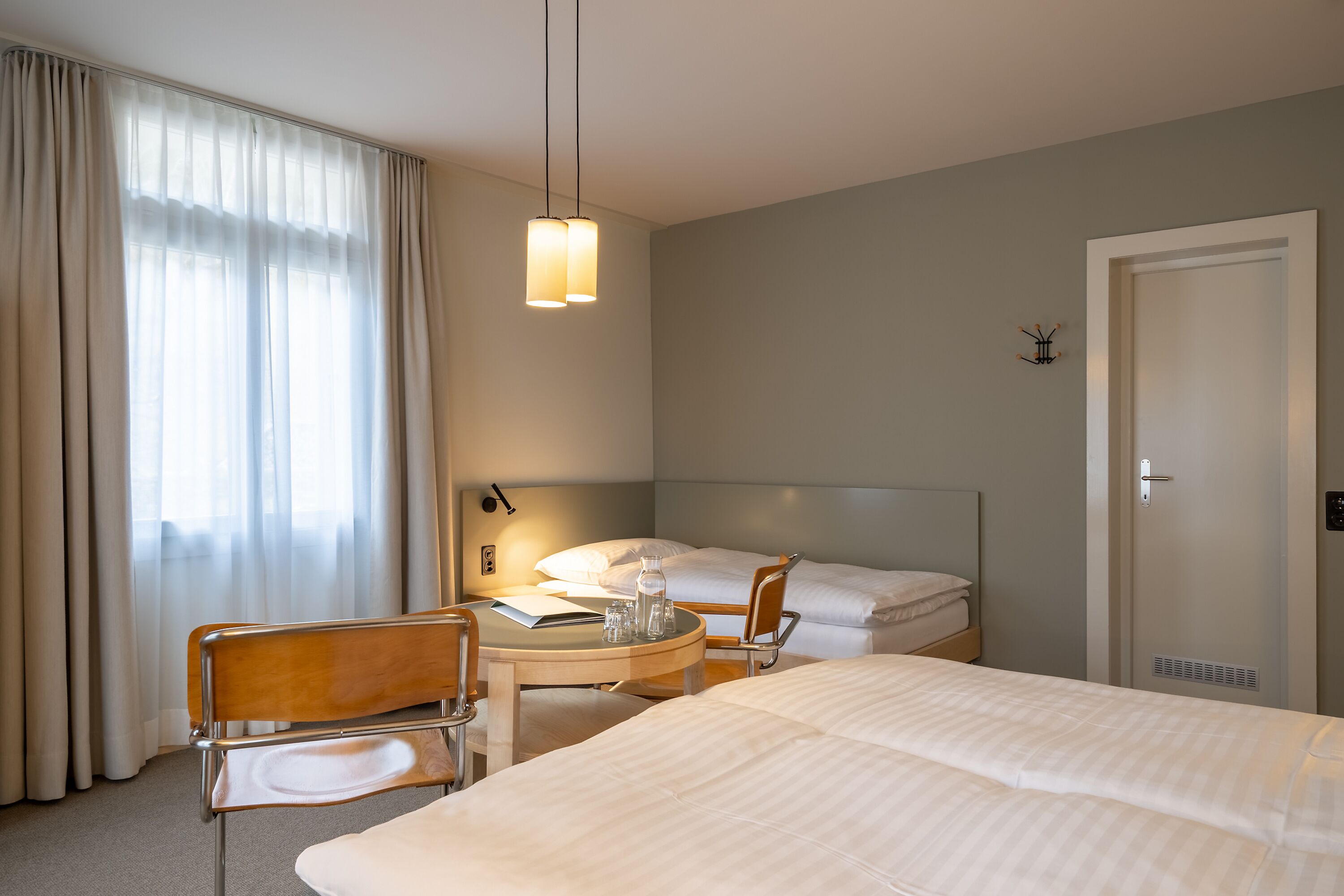 Ein Hotelzimmer mit Bett, Schreibtisch mit Stuhl, Nachttisch mit Lampe und einem Fenster mit Vorhängen.