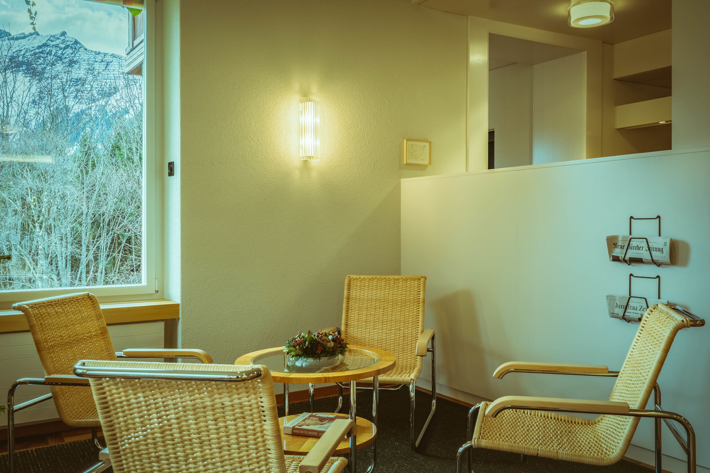 A room with wicker chairs, a round table with a flower vase, a wall-mounted light, and a window with a view of trees.