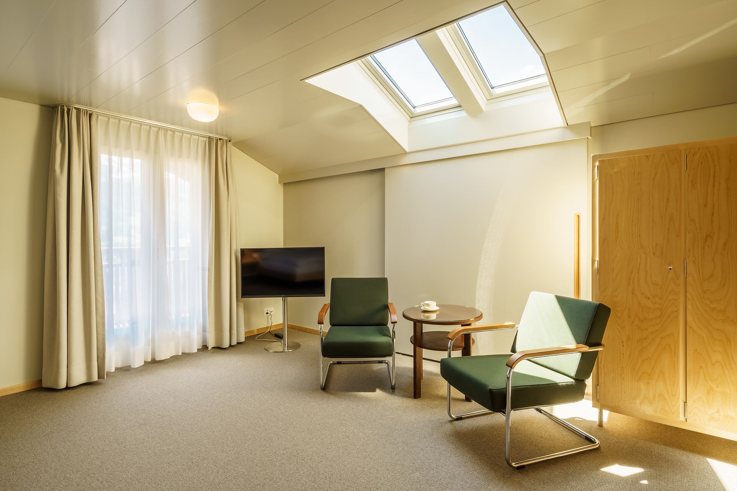 Een hotelkamer met dakraam, televisie, twee groene stoelen, een klein tafeltje en een houten garderobe.