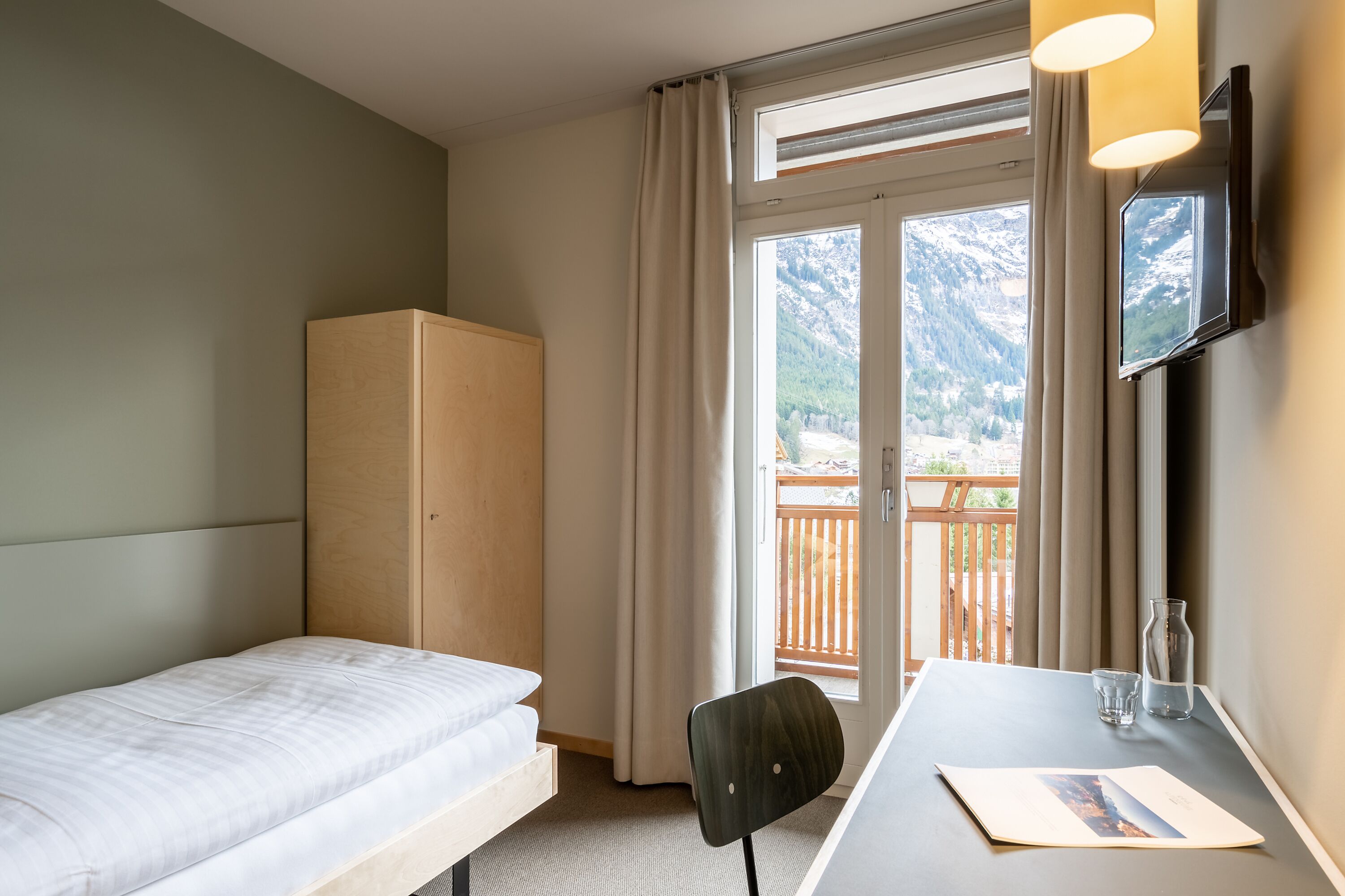 A hotel room with a bed, a desk, a chair, a TV, and a view of mountains through the window.