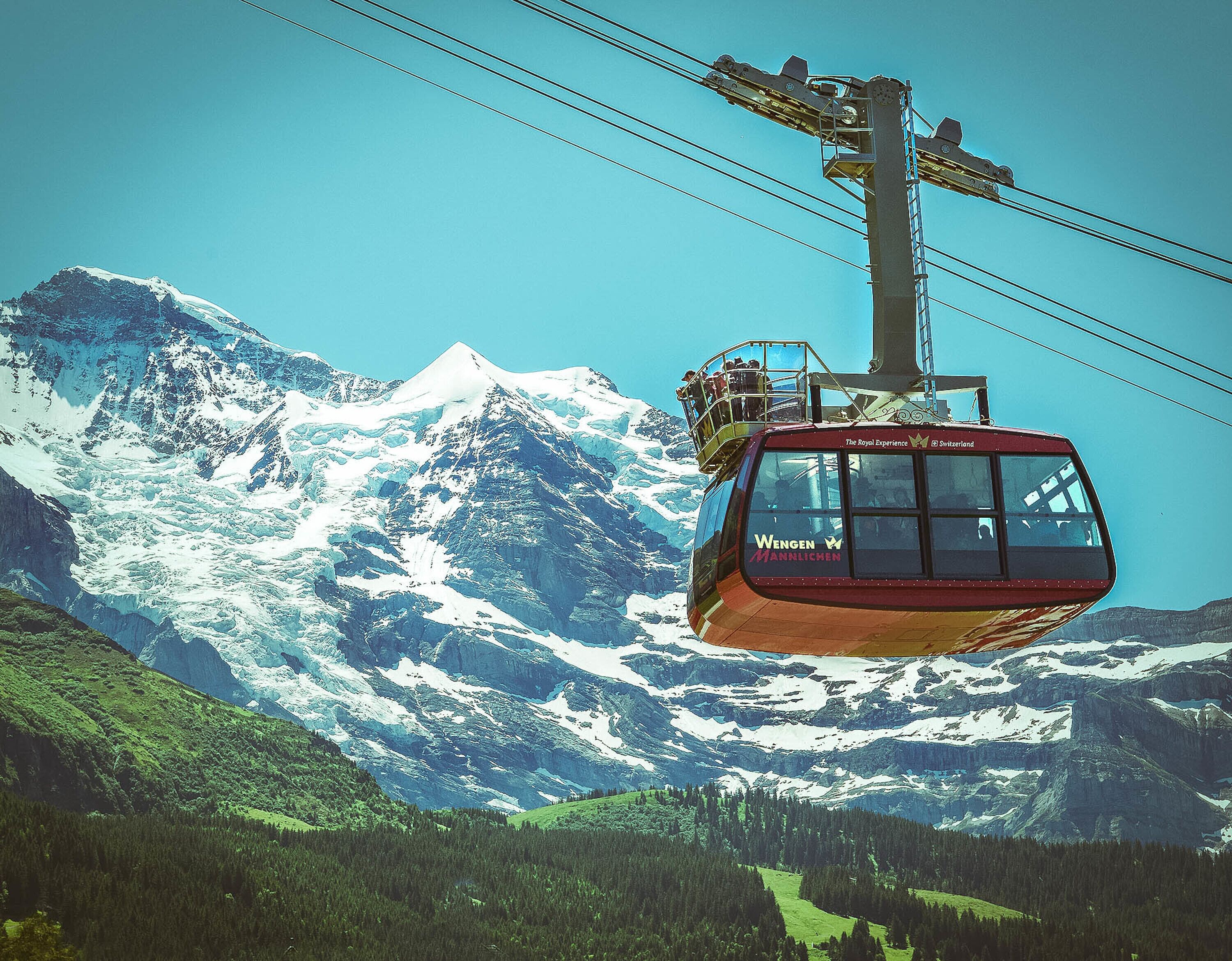 A cable car with "Wengen" and "Männlichen" written on it, moving over a mountainous landscape with snow-covered peaks.