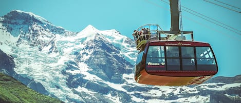 A cable car with "Wengen" and "Männlichen" written on it, moving over a mountainous landscape with snow-covered peaks.
