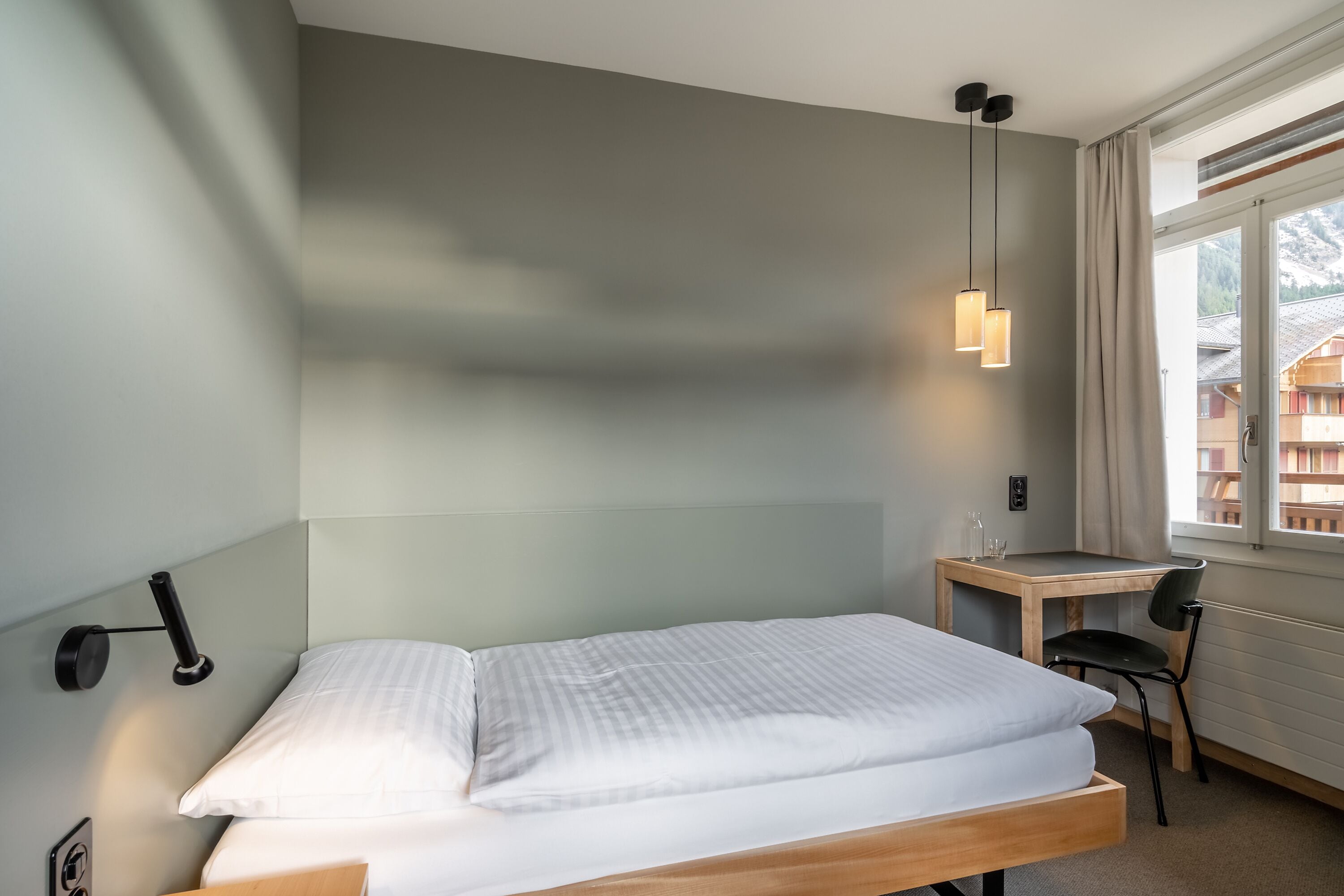 A hotel room with a single bed, a desk, a chair, a lamp, and a window with curtains.