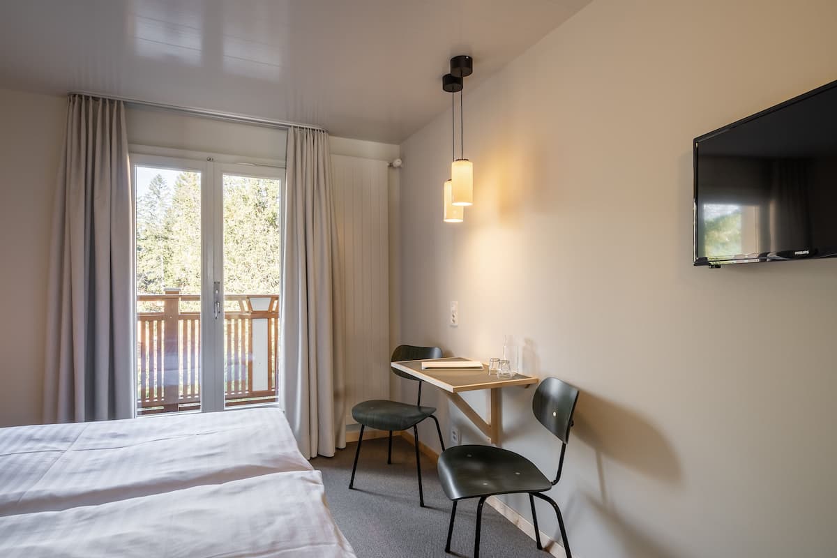 Ein Hotelzimmer mit zwei Betten, einem Schreibtisch mit Stuhl, einem an der Wand montierten Fernseher und einem Balkon mit Vorhängen.