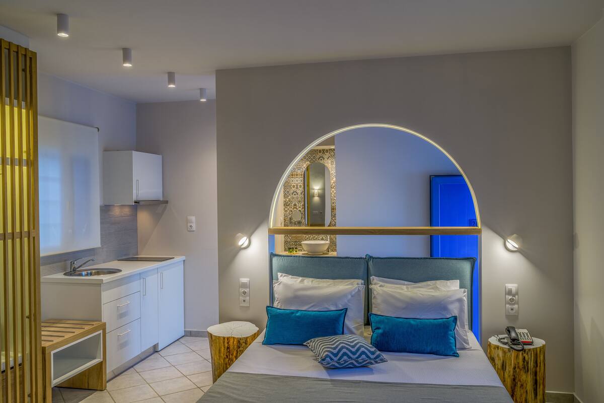 A modern hotel room with a kitchen area, a bed with blue and white bedding, and a unique arched mirror design.