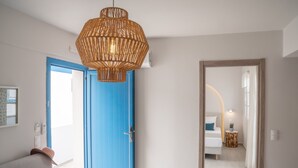 A room with a blue door, a hanging wicker light fixture, a bed with a white headboard, and a small round table with a plant. - Romantica Apartments (Kithira)