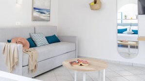 A modern, minimalistic bedroom with a bed, a bedside table, a sofa with cushions, a hanging light fixture, a wall-mounted TV, and a large mirror. - Romantica Apartments (Kithira)