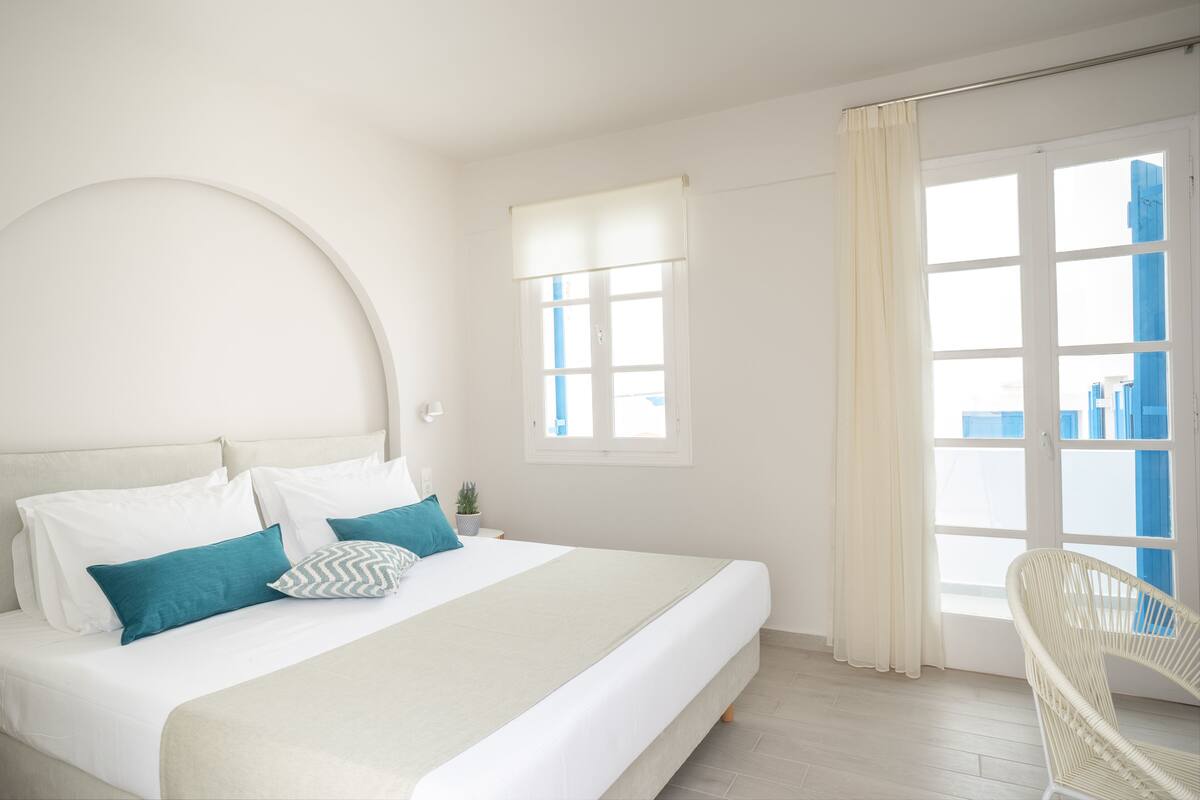 A bedroom with a bed, a chair, a window, and a view of buildings.