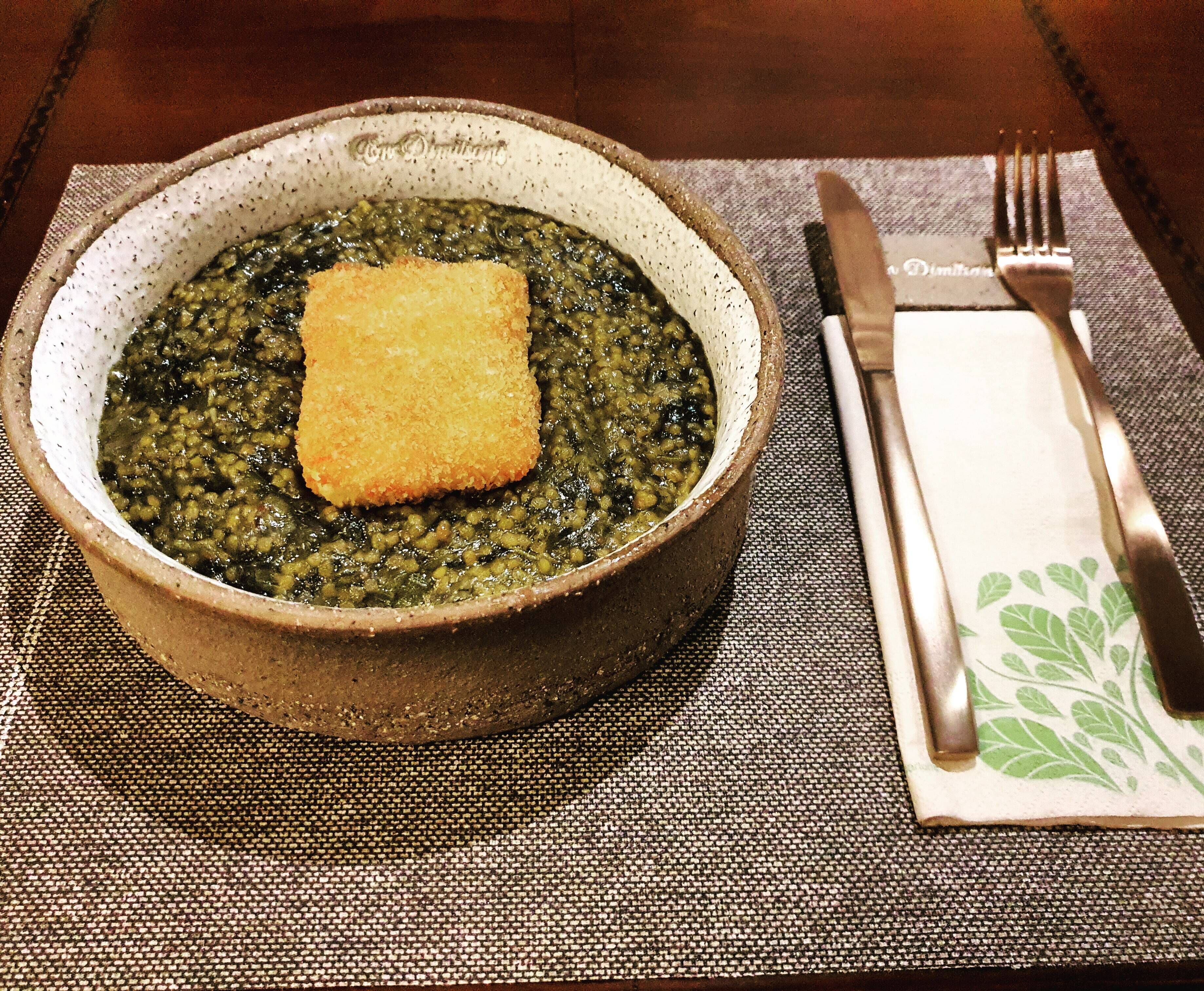 A bowl of soup with a piece of bread on a woven placemat, accompanied by a fork and knife on a napkin.