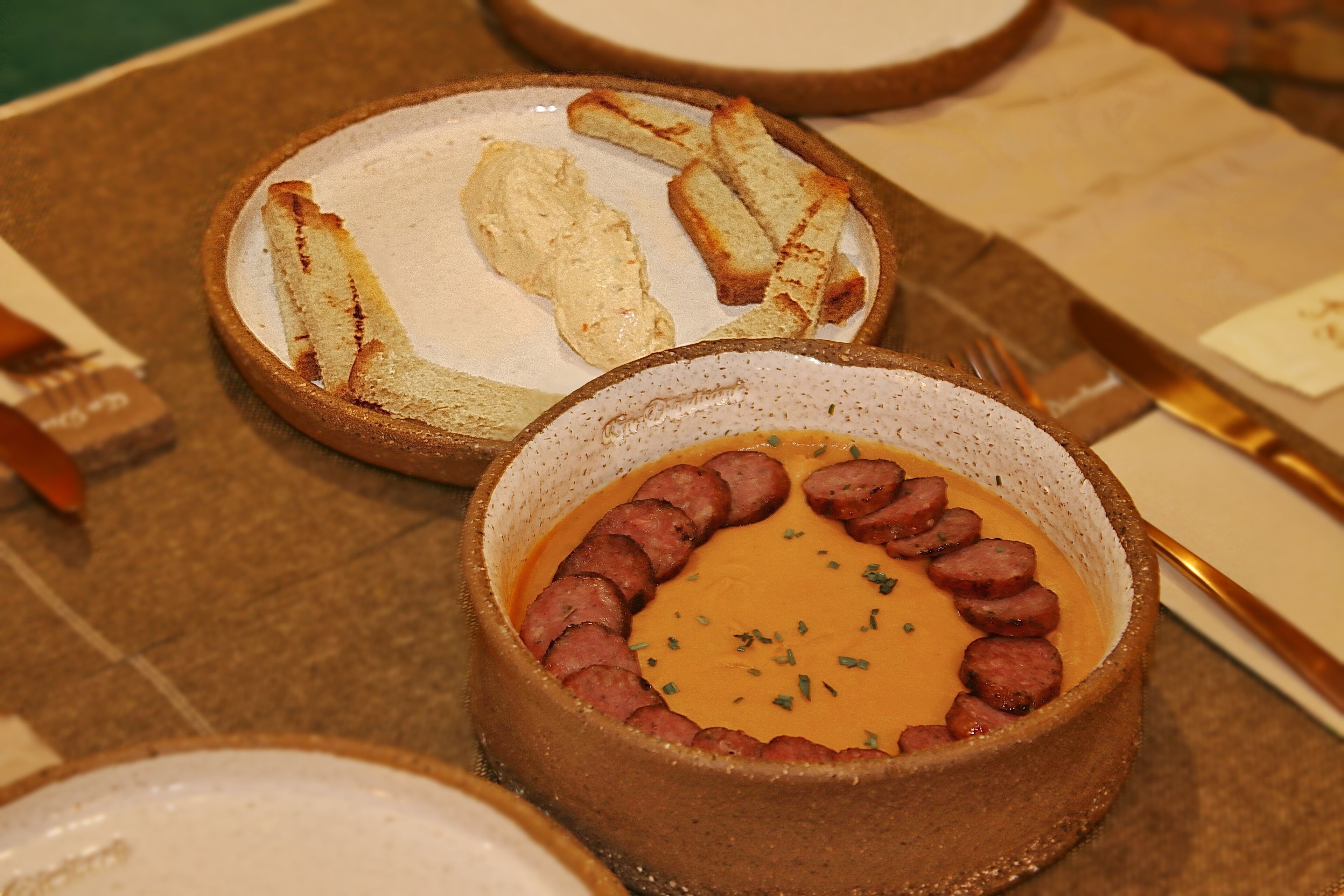 Ein Tisch mit einer Suppenschüssel, garniert mit Wurstscheiben, und einem Teller geröstetem Brot.