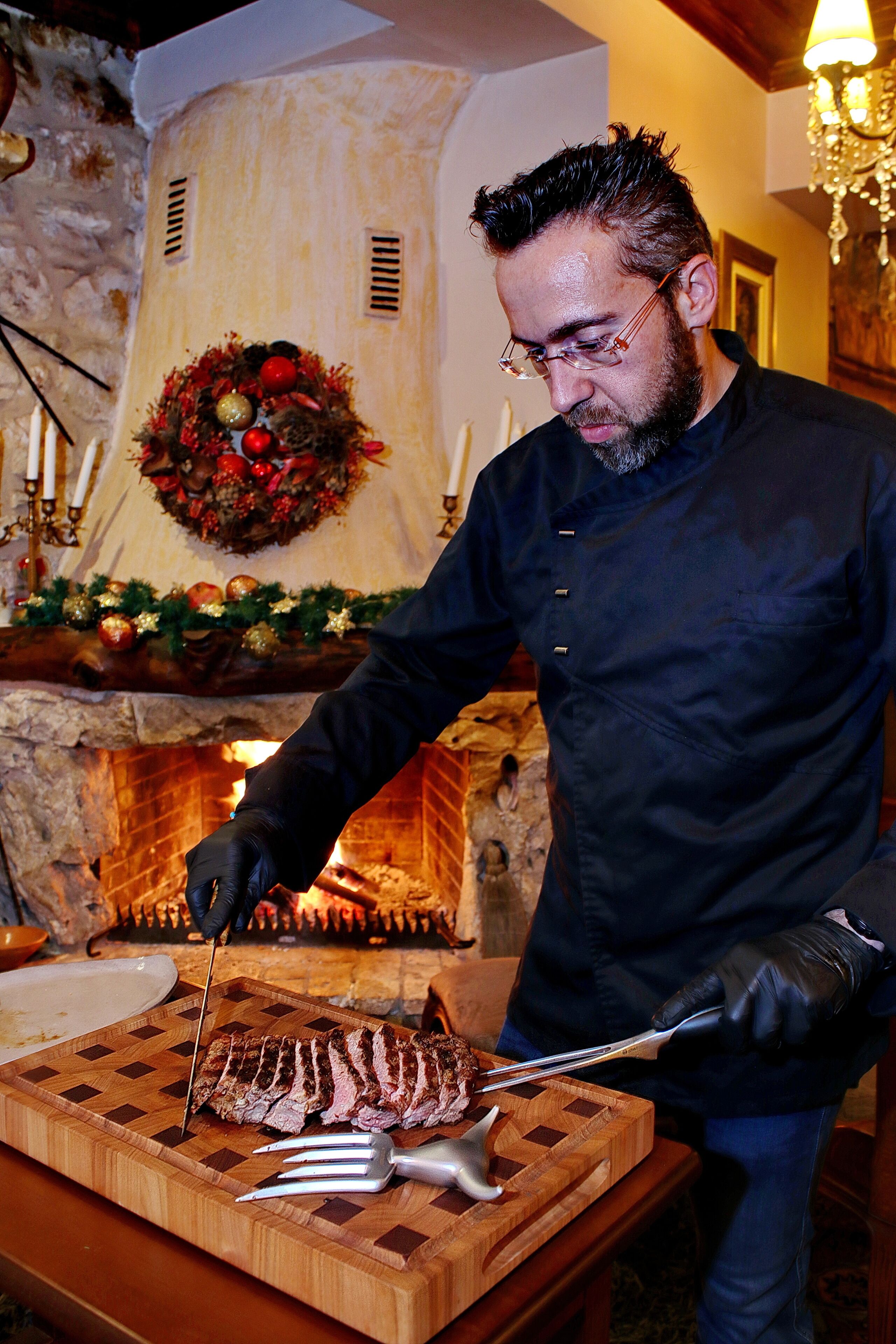 A person slicing meat on a wooden cutting board in front of a fireplace with a Christmas wreath.