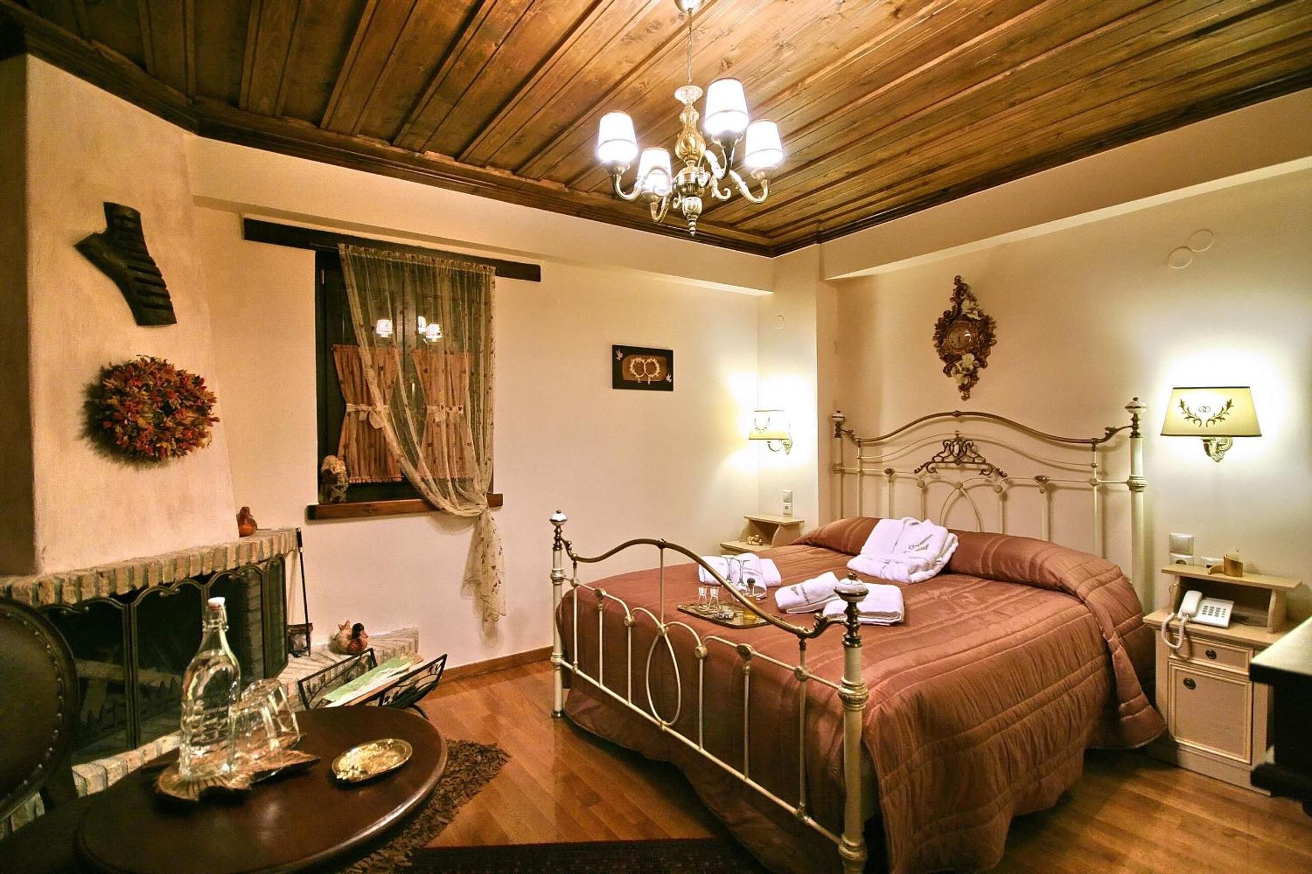 A bedroom with a wooden ceiling, a metal bed frame, a chandelier, a bedside table with a lamp, and a wall-mounted telephone.