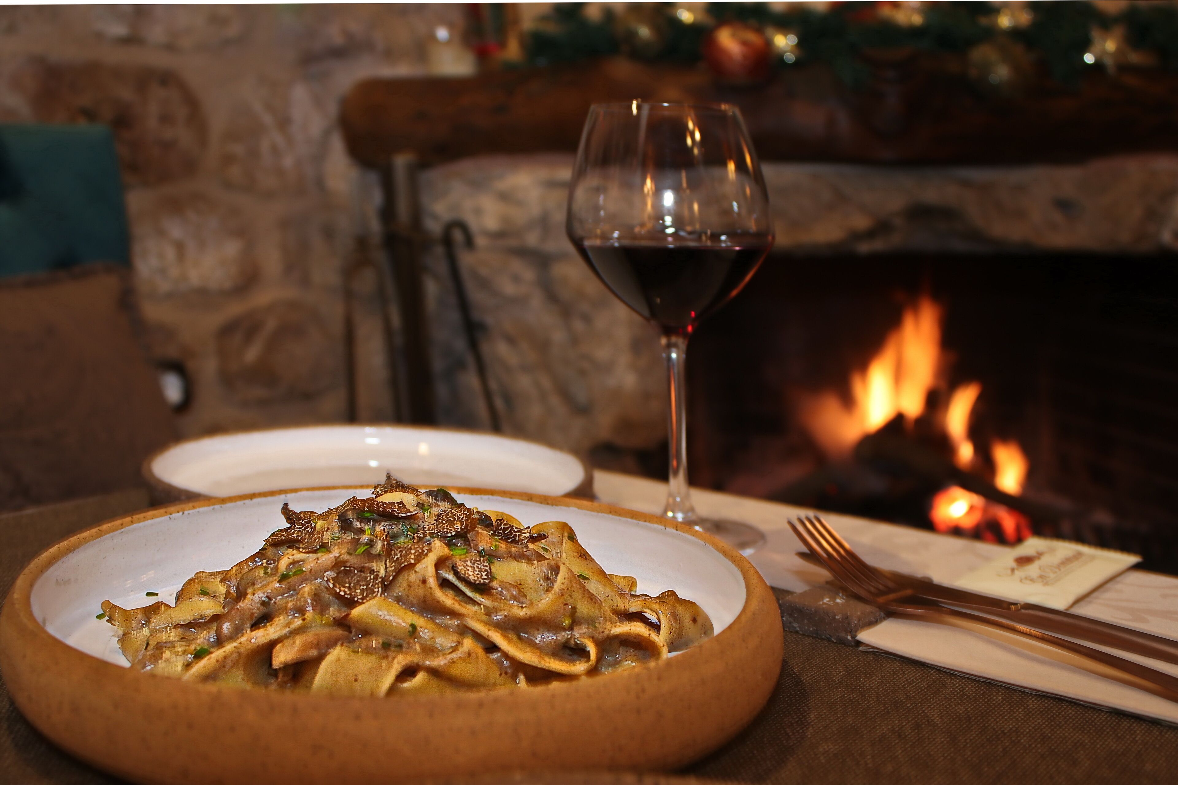 Uma mesa de jantar com um prato de massa, um copo de vinho tinto e uma lareira ao fundo.