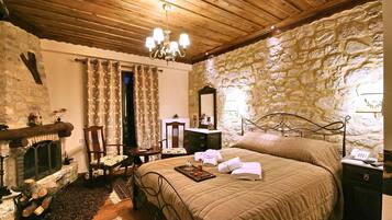 Um quarto aconchegante com parede de pedra, lareira, cama com colcha, mesa de cabeceira e lustre.