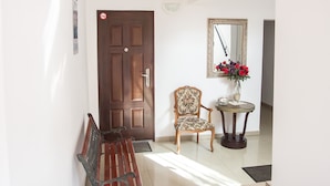 A hallway with a wooden bench, a wall-mounted clock, a door, a chair, a mirror, and a vase with flowers.