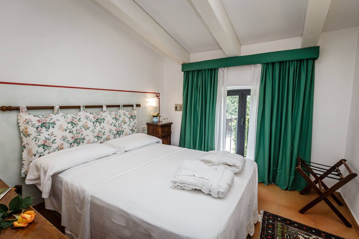 A neatly made bed with white linens and a floral-patterned headboard, a wooden nightstand with a lamp, and a green curtain covering a window.