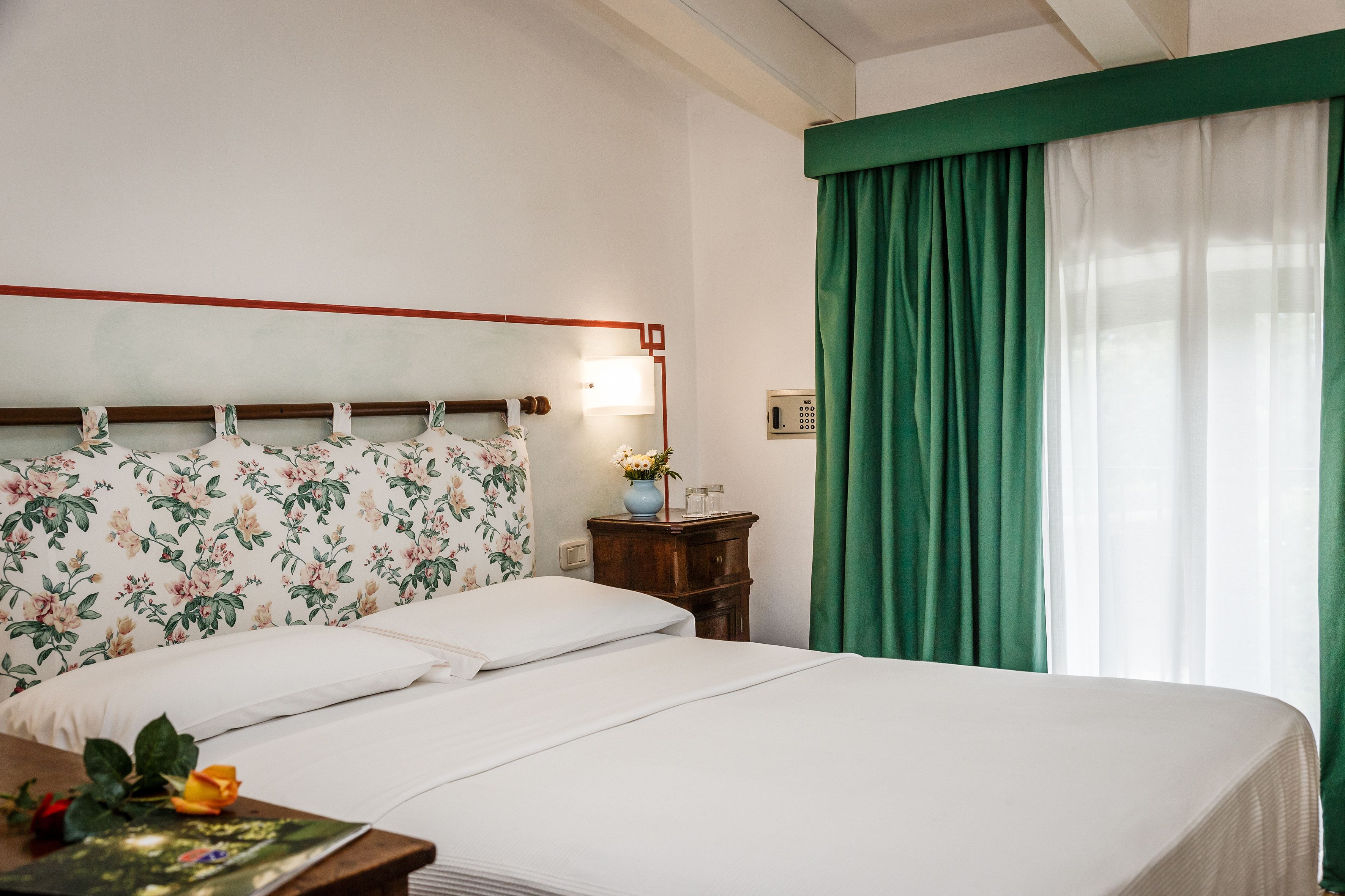 A neatly made bed with floral headboard, a bedside table with a vase of flowers, and a green curtain covering a window.