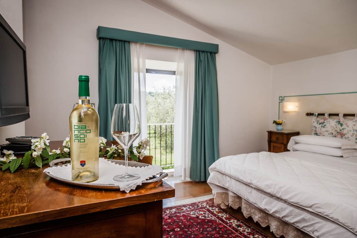 A bedroom with a bed, a bedside table with a bottle and glass, a TV, and a window with curtains.