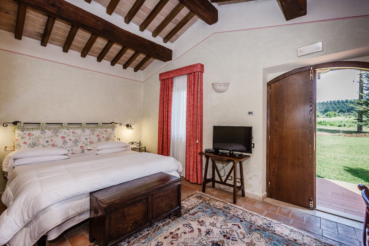 A bedroom with a bed, a television, a wooden bench, a rug, and a door leading to an outdoor area.