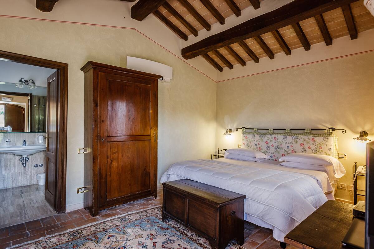 A bedroom with a wooden wardrobe, a bed with white bedding, a bench, and a bathroom visible through an open door.