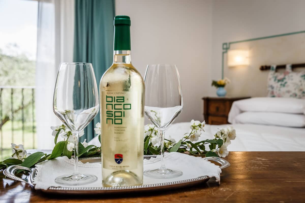 A bottle of white wine on a tray with two glasses, a bed, and a nightstand with a lamp in a hotel room.