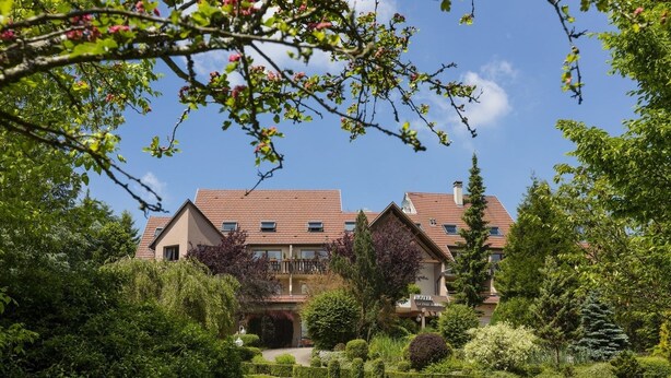 A hotel with a traditional European architectural style, featuring a symmetrical facade, multiple windows, and a well-maintained garden with various shrubs and a lawn. - Le Petit Kohlberg (Lucelle)