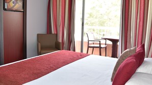 Une chambre d’hôtel avec un lit, une chaise, un balcon avec des rideaux et un téléviseur fixé au mur.