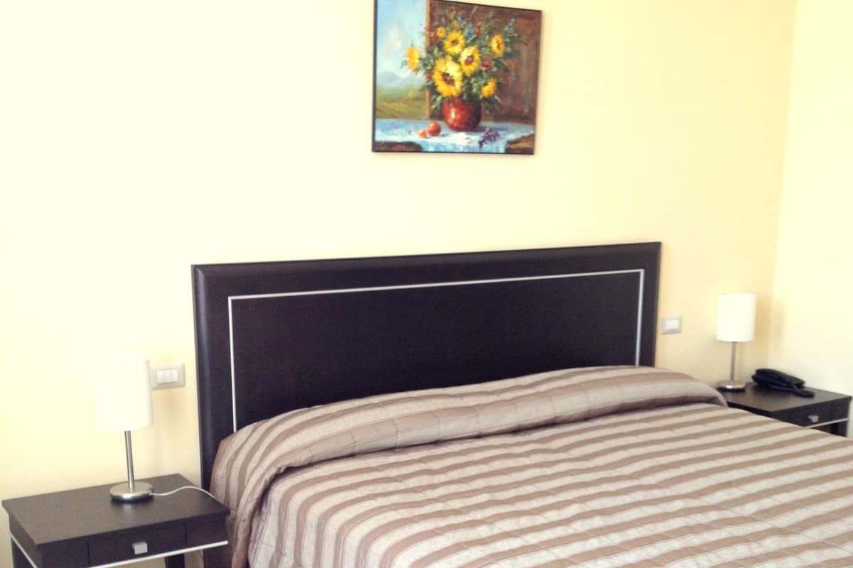 Hotelkamer met een bed, nachtkastje, lamp, schilderij van zonnebloemen en een houten vloer.