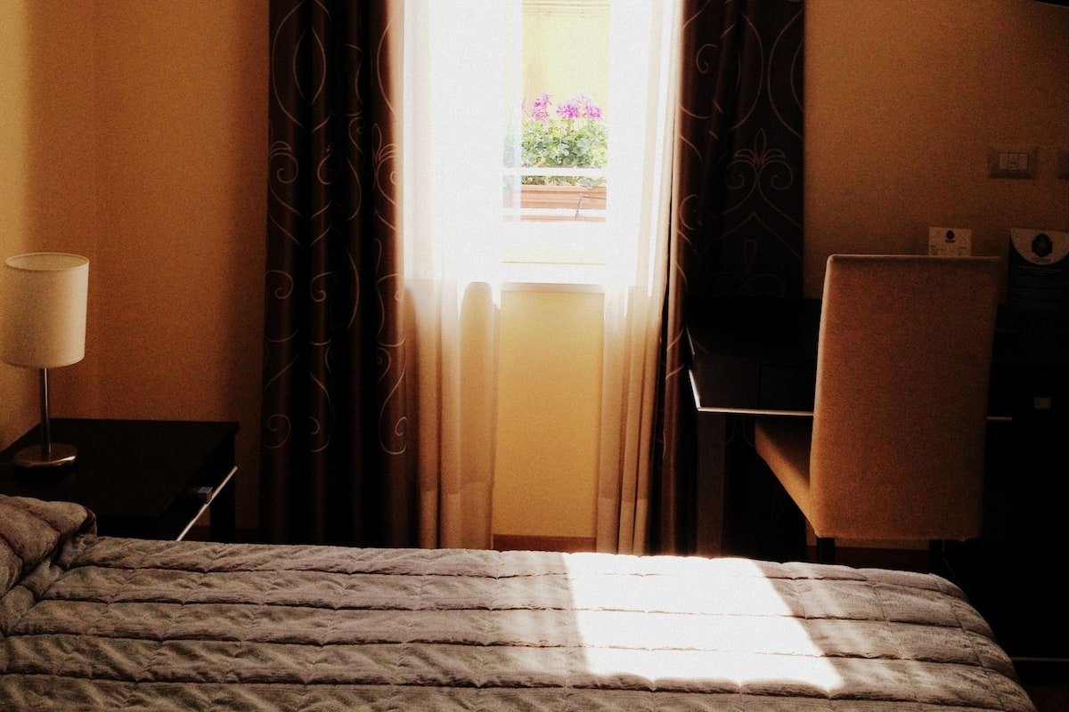 Een hotelkamer met een bed, een bureau, een stoel, een lamp en een raam met gordijnen.