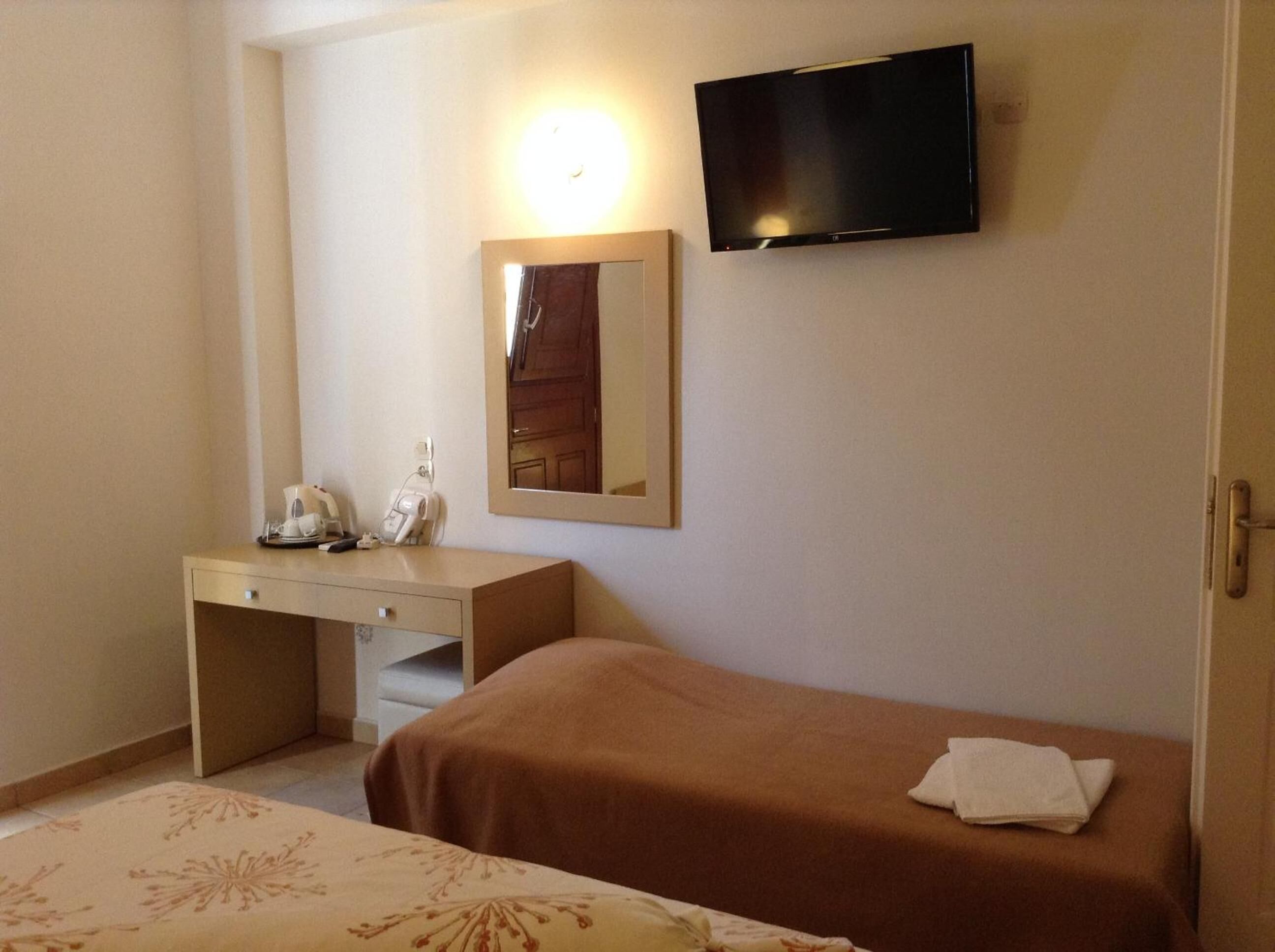 A hotel room with a single bed, a bedside table with a telephone and a coffee maker, a flat-screen TV mounted on the wall, and a mirror above the table.