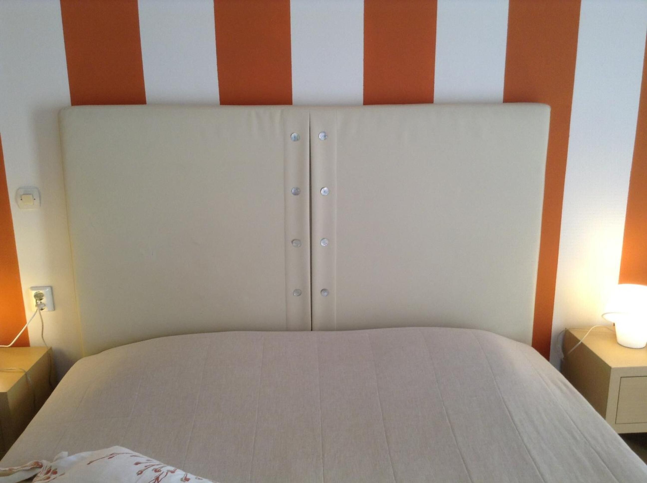 Een bed met een beige hoofdbord en twee nachtkastjes, waarvan er één een lamp heeft.