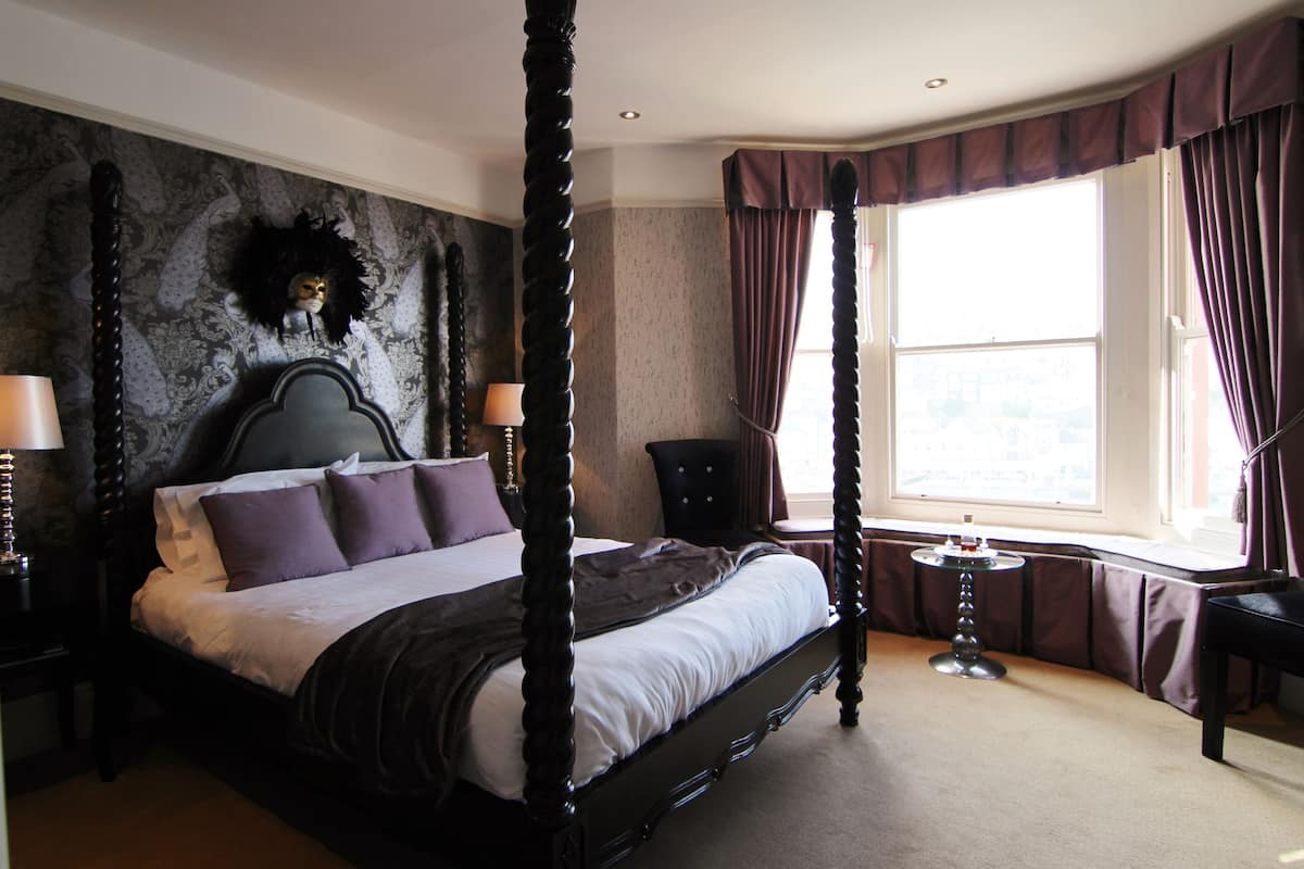 A four-poster bed with a dark headboard, a round table, and a view of the city through a large window with curtains.