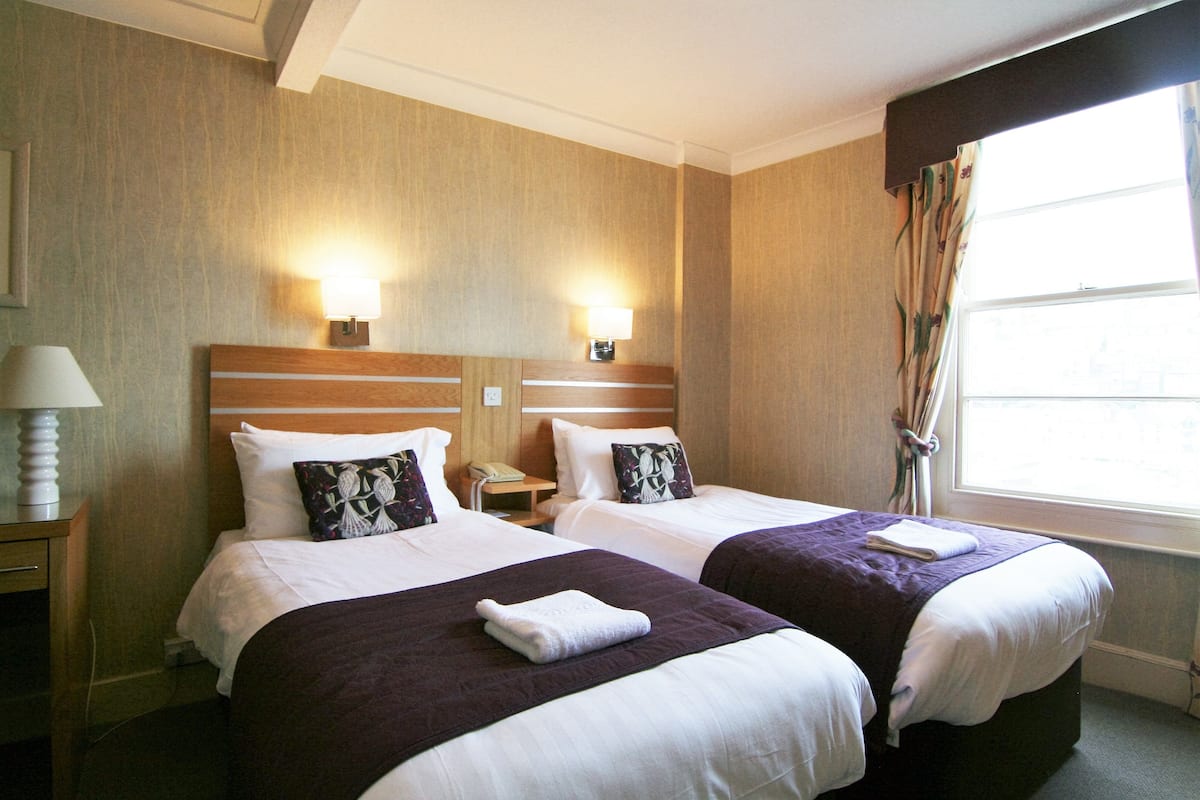 A hotel room with two beds, each with a purple bedspread and white linens. There are two wall-mounted lamps on either side of the beds.